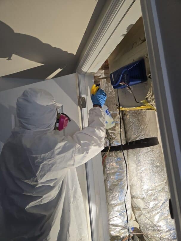 A technician in protective gear applying mold remediation treatment to the area around a doorframe in a home with visible ductwork.